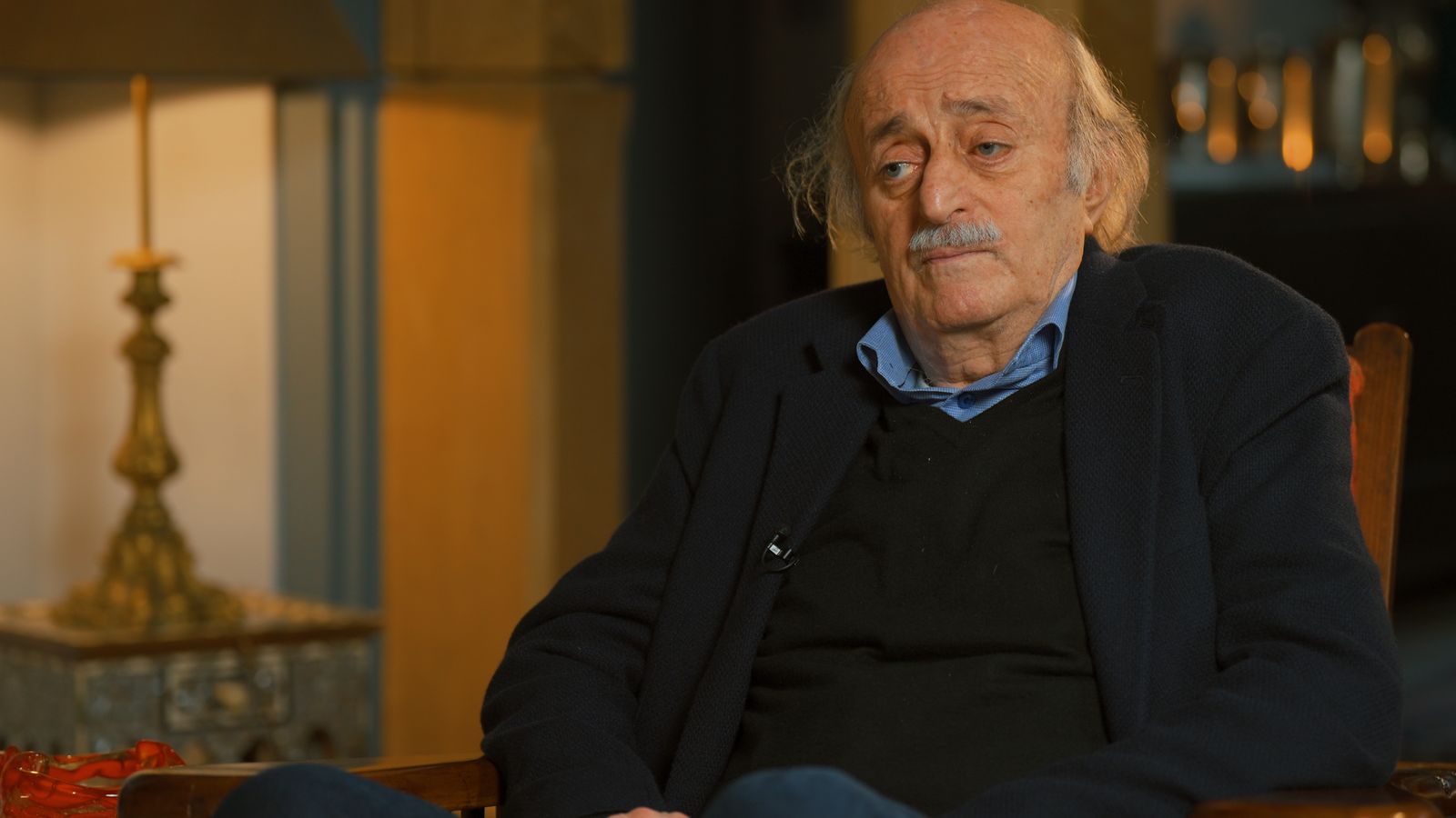 A seated interview lit on location: warm practical lamps, portraits and bookshelves held in frame behind the subject